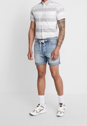 Mann trägt ein kurzärmeliges weiß-grau gestreiftes Hemd, hellblaue Jeansshorts mit gebundenem weißem Kordelzug, weiße Socken und weiße Turnschuhe.