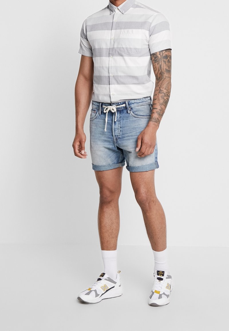 Homme portant une chemise à manches courtes blanche et grise à rayures, un short en jean bleu clair avec un cordon blanc noué, des chaussettes blanches et des baskets blanches.