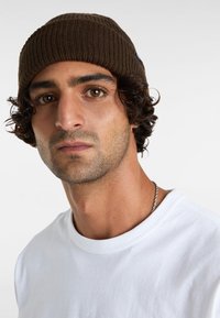 Braune gerippte Mütze mit engem Sitz, aus Strickmaterial gefertigt, getragen von einer Person in einem weißen T-Shirt, das eine dezente metallische Kette aufweist.