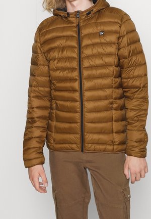 Person trägt eine braune gesteppte Kapuzenjacke mit Reißverschluss, kombiniert mit braunen Cargohosen, und steht vor einem schlichten Hintergrund.