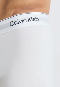Krupan plan osobe koja nosi bijelo donje rublje marke Calvin Klein s vidljivim logotipom na pojasu, na neutralnoj pozadini.
