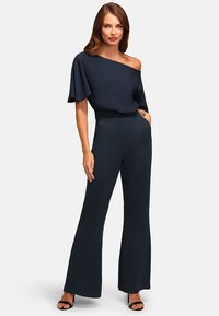 Maritieme one-shoulder jumpsuit met wijde pijpen. Zacht materiaal, uitlopende ontwerp en pocketdetails, gecombineerd met zwarte open-toe hakken.