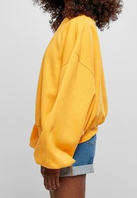 Sudadera amarilla de gran tamaño con hombros caídos, puños acanalados y una textura suave, combinada con shorts de mezclilla.