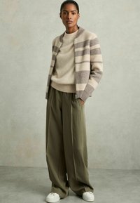 Texturerad beige och brun randig cardigan över en beige tröja, tillsammans med lösa olivgröna byxor och vita sneakers. Neutral färgpalett.