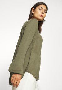 Olive green long-sleeve top with a relaxed fit, textured fabric, and gathered cuffs. Hemline is slightly longer at the back.
