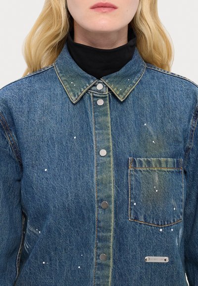 Jachetă din denim albastru cu un buzunar frontal, accesorii din metal argintiu și detalii cu stropi de vopsea albă. Prezintă un guler clasic și închidere cu nasturi.