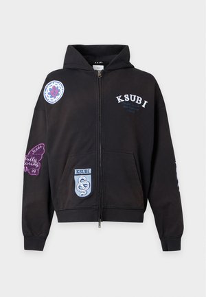 Homme portant une casquette noire, un sweat à capuche noir avec des écussons, une chemise blanche, un pantalon de survêtement noir et des baskets noires et blanches, debout devant un fond blanc.