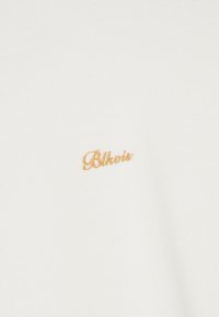Tessuto a maglia bianco con un piccolo logo ricamato in oro "Blkivs" sul lato sinistro. La texture è liscia e uniforme.