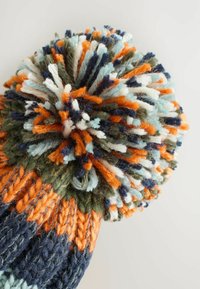 Bonnet tricoté avec un pompon multicolore sur le dessus. Les couleurs incluent l'orange, le bleu, le vert et le blanc. Fil texturé avec des motifs entrelacés.