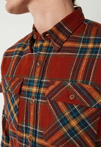 Chemise à carreaux rouge et bleue avec col boutonné, poches poitrine, boutons marron et tissu en flanelle texturé.