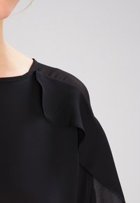 Černá blůza s hladkým materiálem a jemným drapovaným designem, průsvitnými rukávy a kulatým výstřihem se volánkovým akcentem na rameni.