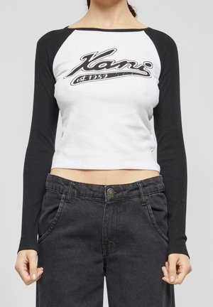 Kvinde iført en hvid og sort langærmet crop top med teksten "Kani est. 1989" og høj talje mørkegrå jeans.