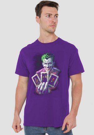 Man wearing purple t-shirt featuring Joker with green hair holding four Joker playing cards, looking slightly to the side.
