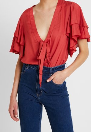 Kobieta ubrana w czerwoną bluzkę z krótkim rękawem, ozdobioną falbanami, z głębokim dekoltem w serek i wiązaniem z przodu, w połączeniu z wysokim stanem ciemnoniebieskich dżinsów, z ręką w kieszeni.