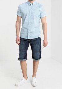 Camisa de manga corta azul claro con un pequeño diseño estampado, combinada con pantalones cortos de mezclilla oscura y zapatillas blancas.