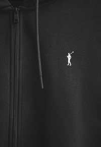 Sudadera con cremallera negra hecha de una tela suave, que presenta un pequeño logo bordado en blanco de una figura con un palo de golf en el lado izquierdo del pecho.