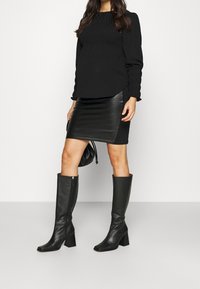 Černá blůza s dlouhým rukávem a nařasenou texturou, zkombinovaná s černou koženou mini sukni a vysokými koženými botami na podpatku, vystihující elegantní design.