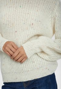 Helles, cremefarbenes Strickpullover mit lockerer Passform, versehen mit bunten Sprenkeln, gerippten Bündchen und einem strukturierten Gewebe, kombiniert mit dunkelblauen Jeans.