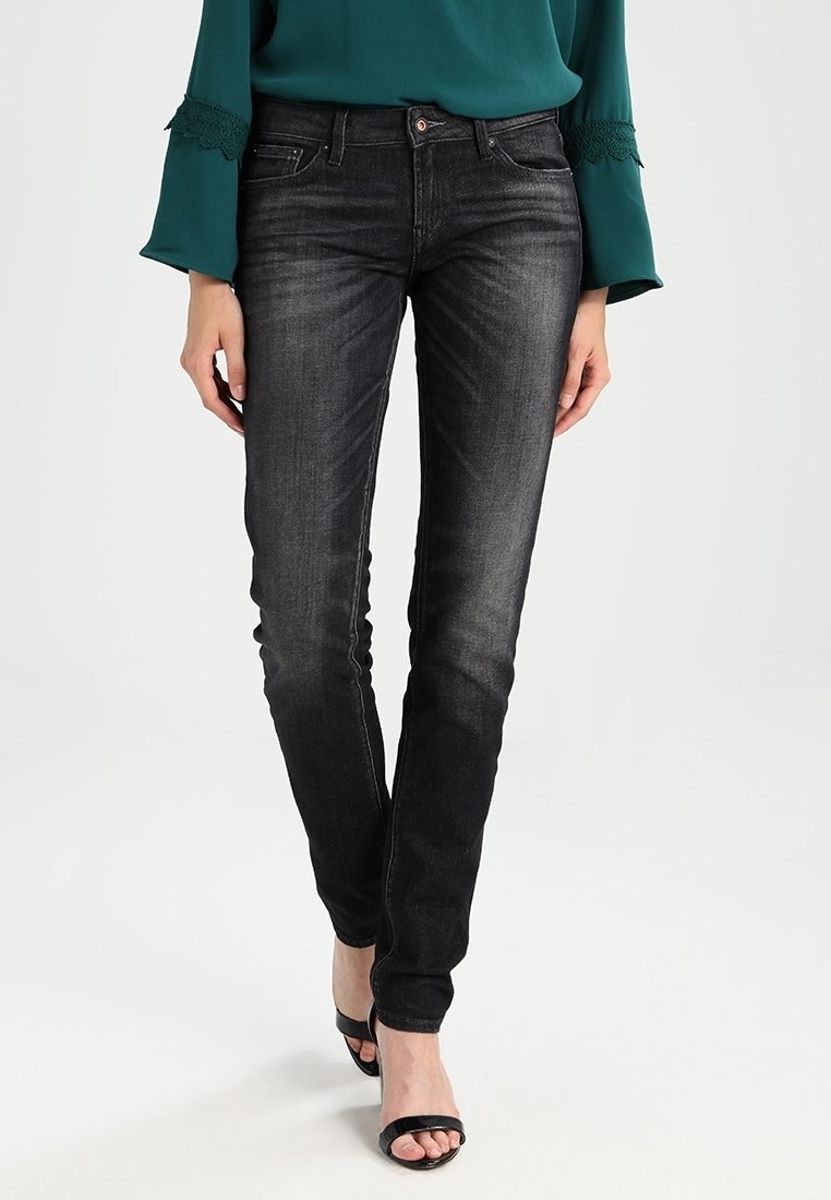 Jean skinny noir en denim. Présente une texture légèrement délavée, cinq poches et une coupe ajustée. Porté avec des talons noirs.