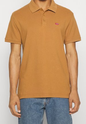 Brązowa bawełniana koszulka polo z kołnierzykiem, z trzema guzikami oraz małym czerwonym logiem Levi's na lewej piersi. W zestawie z niebieskimi dżinsami.