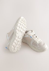 Viegls skrienamais apavi bēšā režģa materiālā ar atstarojošiem akcentiem, teksturētu gumijas zolīti un gaiši zilu detaļu sānos.