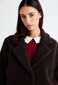 Manteau en fausse fourrure marron avec un col cranté, présentant une surface texturée, superposé à un pull rouge avec un col blanc en dessous.
