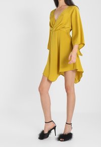 Robe enveloppante jaune avec un décolleté en V profond, des manches longues et un ourlet asymétrique. Portée avec des sandales à talons hauts noires ornées de détails en fourrure.
