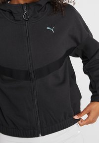 Veste de sport noire zippée avec capuche, bande texturée contrastante et petit logo turquoise sur la poitrine. Poignets et taille élastiques.