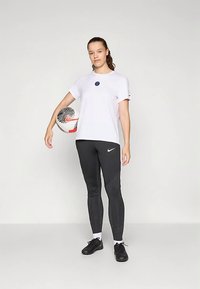 White short-sleeve t-shirt with a circular blue logo, paired with black textured leggings and black soccer shoes, holding a soccer ball.