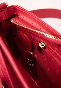 Interno di una borsa a tracolla rossa in pelle con una texture liscia, dotato di una tasca con zip, accessori dorati e un dettaglio circolare con logo impresso.