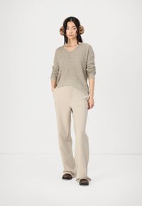 Maglione lavorato a maglia beige con scollo a V e texture a costine, abbinato a pantaloni beige a gamba larga. Indossatore con cuffie in pelliccia maculata; posa eretta.