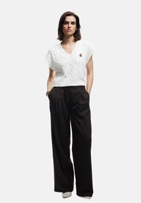 Hvid strikket sweater med kabeldesign og V-hals, parret med højtaljede sorte bukser. Bemærkelsesværdigt broderet bjørnelogo på sweateren.
