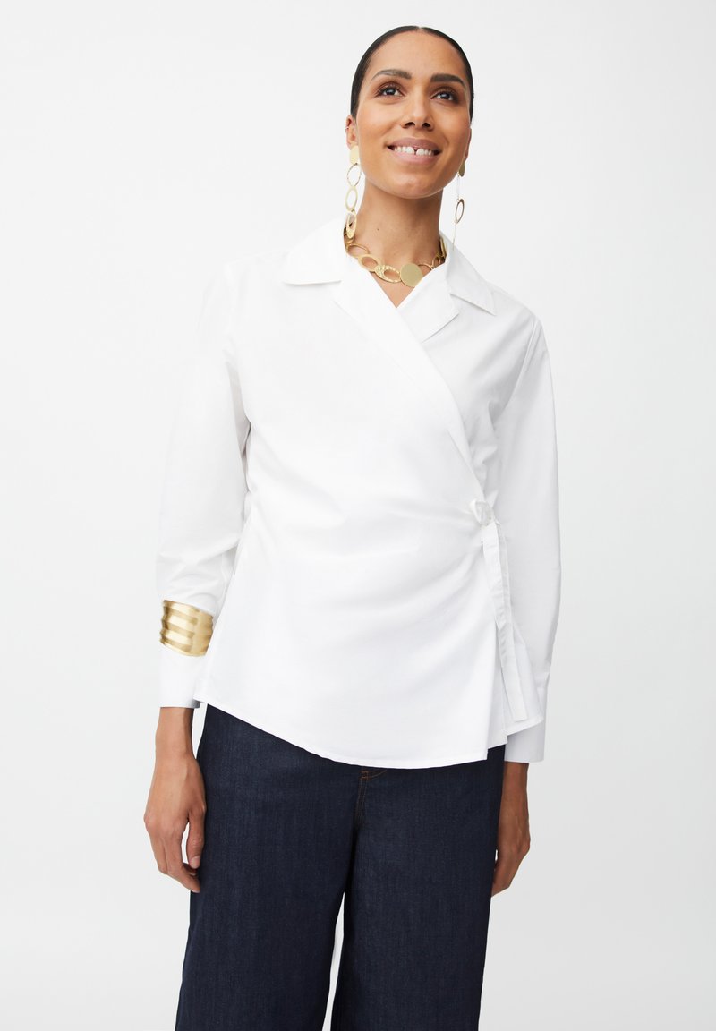 Camisa blanca con cuello en pico, detalles de hardware dorado texturizado en las mangas y un diseño ceñido para un ajuste entallado. Combinada con jeans oscuros.
