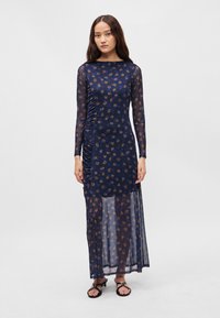 Námořnické šaty s dlouhým rukávem, květinovým vzorem, řasením na jedné straně, průhlednou tkaninou a rovnou siluetou, doplněné černými sandály.