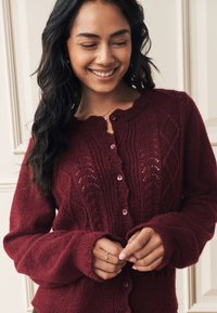 Bordeauxfarbener, gestrickter Cardigan mit einem wellenförmigen Ausschnitt, Knopfleiste vorne und strukturierten Rautenmuster. Lange Ärmel mit gerippten Bündchen.