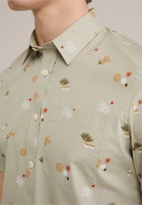 Krátký rukáv, světl zelená košile s vícero barevnými vzory, které zahrnují rostliny, vlnky a abstraktní tvary. Obsahuje náprsní kapsu a knoflíkovou légou.