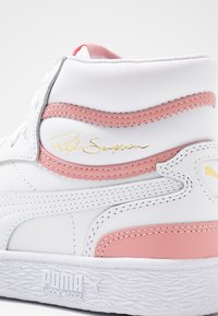 Bílé vysoké tenisky vyrobené z kůže, s růžovými akcenty a zlatým podpisovým detailem. Strukturální bílá gumová podrážka se značkováním.