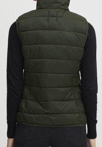 Zielona kamizelka puchowa z wysokim kołnierzem, z poziomymi przeszyciami, pikowaną fakturą i dopasowanym krojem. Nosi założoną na ciemny sweter.