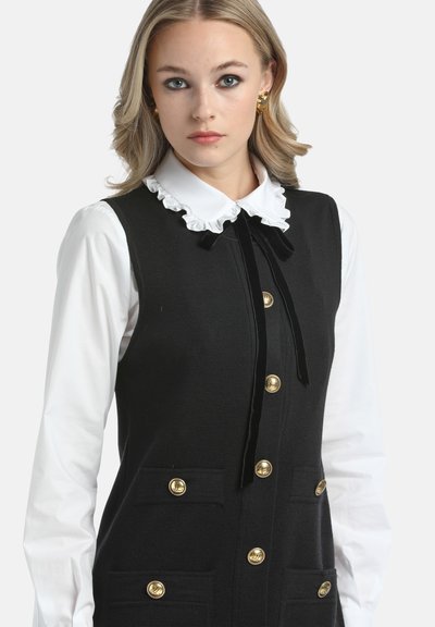 Abito nero lavorato a maglia con bottoni in oro e tasche, dotato di un colletto bianco a balze e un nastro nero. Tessuto con design testurizzato.