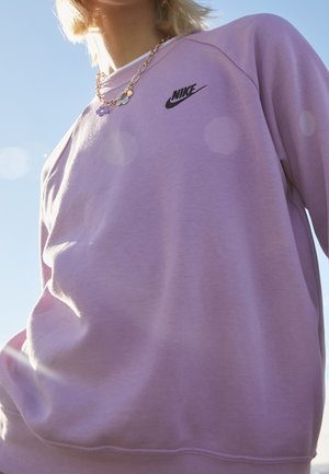 Osoba nosząca jasnofioletową bluzę Nike i złoty łańcuszek z zawieszkami w kształcie kwiatów na tle czystego niebieskiego nieba.
