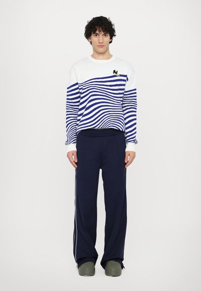 Marineblaue Sweatpants und ein weißer Strickpullover mit blauen Wellenmustern. Der Pullover hat ein gelbes Logo-Detail auf der Brust.