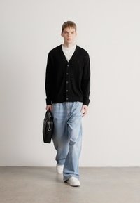 Casaco em malha preto, T-shirt branca, jeans azul claro de perna larga, mala preta com ferragens prateadas, tênis brancos, fundo neutro.