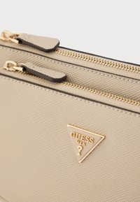 Bézs színű, texturált cipzáras tok arany színű fém elemekkel, két cipzáras rekesszel és egy háromszög alakú, "GUESS USA" feliratú logóval.