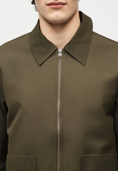 Homme portant une veste vert olive avec col montant et fermeture éclair argentée à l'avant, vue partielle des épaules au menton.