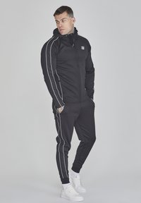 Schwarzer sportlicher Trainingsanzug aus einem glatten, dehnbaren Material mit weißen seitlichen Streifen. Enthält eine zipfelbare Kapuzenjacke und enge Hosen. Weiße Sneakers.