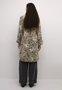 Duga majica s cvjetnim uzorkom u bež boji s zelenim i plavim naglascima, s naglašenim rukavima i slobodnim krojem. Uspođena s hlačama širokih nogavica u sivoj boji.