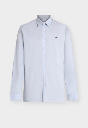 Ανοιχτό μπλε πουκάμισο με μακριά μανίκια και κουμπιά, με μυτερό γιακά και πράσινο λογότυπο κροκοδείλου στο αριστερό στήθος, σε λευκό φόντο.