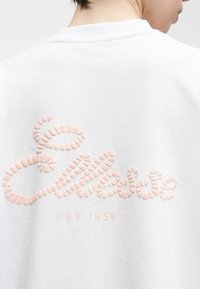 Άτομο που φοράει λευκό φούτερ με ανάγλυφο, υφασμάτινο κείμενο "Esprit" και "EST 1959" τυπωμένα στην πλάτη σε απαλό ροζ χρώμα.