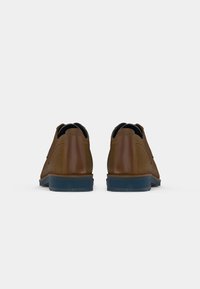 Um par de sapatos masculinos em couro castanho e camurça, vistos de trás, sobre um fundo branco liso.