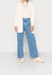 Kerut sweater krem, kemeja bergaris putih dan abu-abu, jeans biru dengan pinggang tinggi, dan sepatu bot chunky beige. Menampilkan siluet kaki lebar dan aksen berkawat.